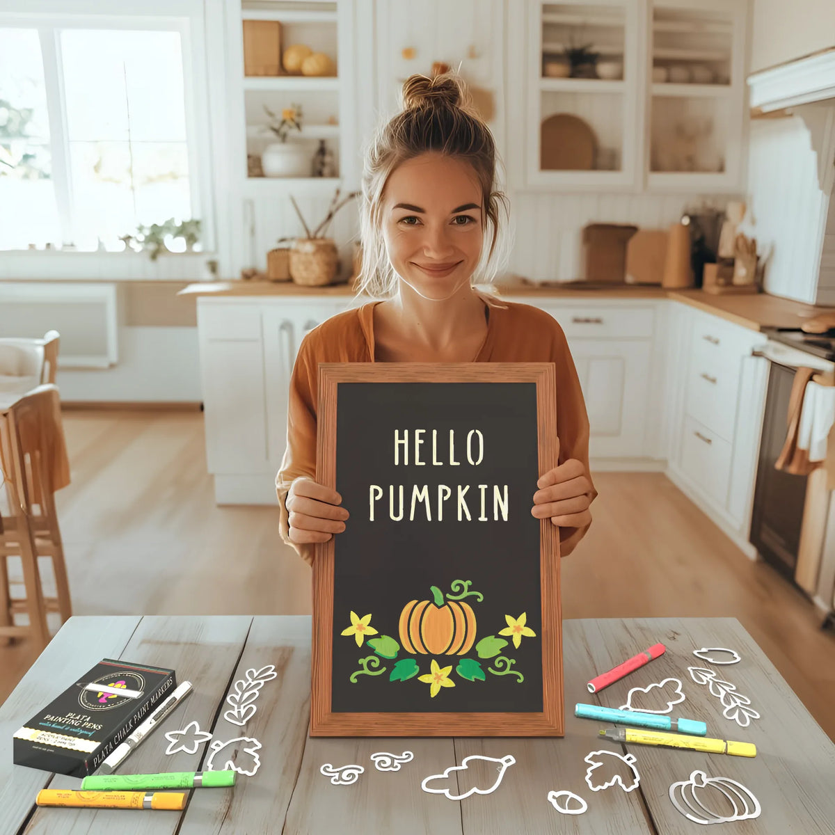 A woman holding a framed chalkboard in a cozy farmhouse kitchen, featuring fall chalkboard art. The chalkboard displays the message &#39;Hello Pumpkin&#39; along with a pumpkin chalk art created using Plata Chalkboards&#39; fall stencils and colorful chalk markers to create a DIY fall decor.  Fall leaf and acorn stencils and markers are scattered on the table beside her