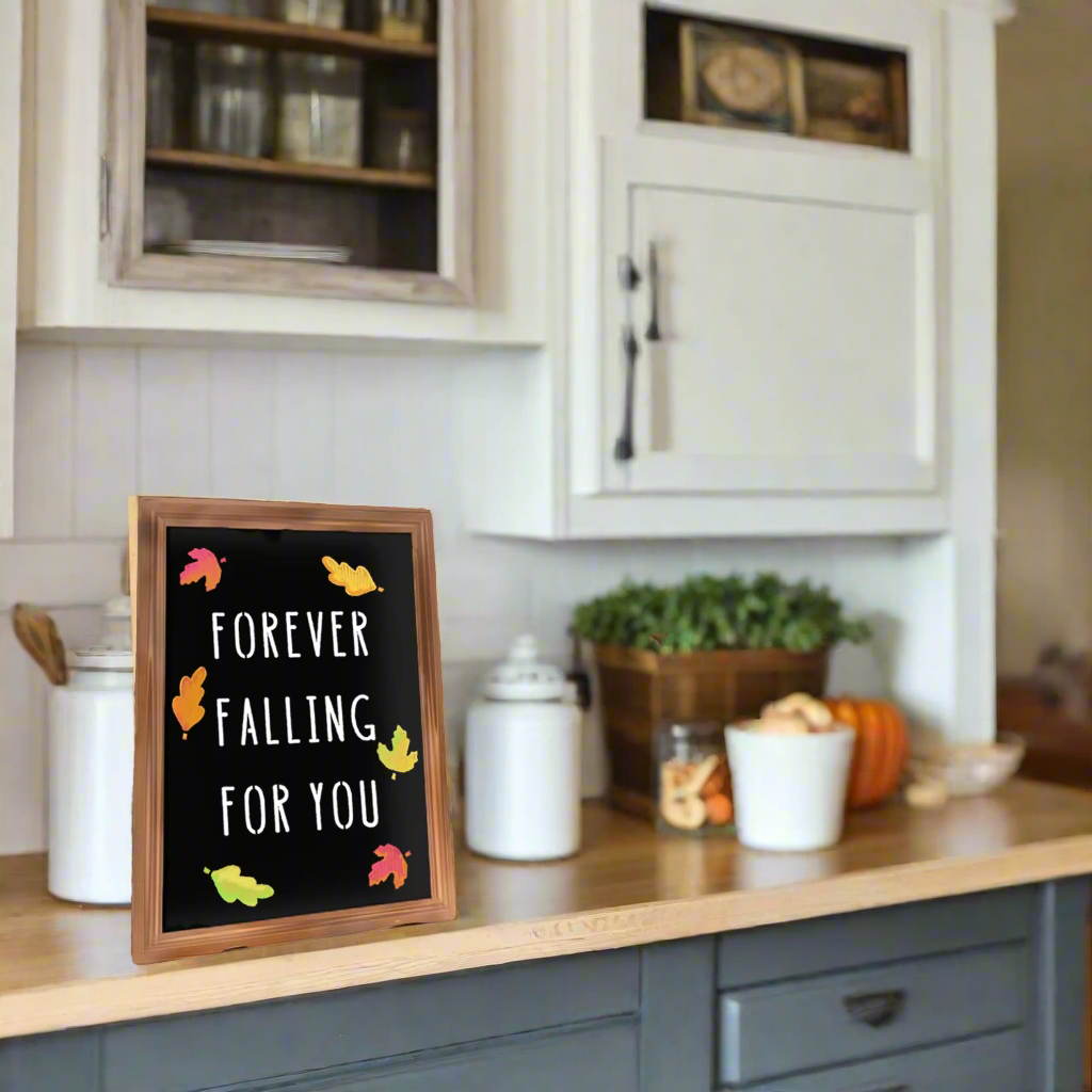 A bride and groom share a romantic moment at their rustic fall wedding, with a framed fall wedding chalkboard in the foreground. The chalkboard features the message &#39;Forever Falling For You&#39; along with autumn leaf designs, crafted using chalkboard stencils from Plata Chalkboards. Lanterns with string lights enhance the cozy, rustic atmosphere.